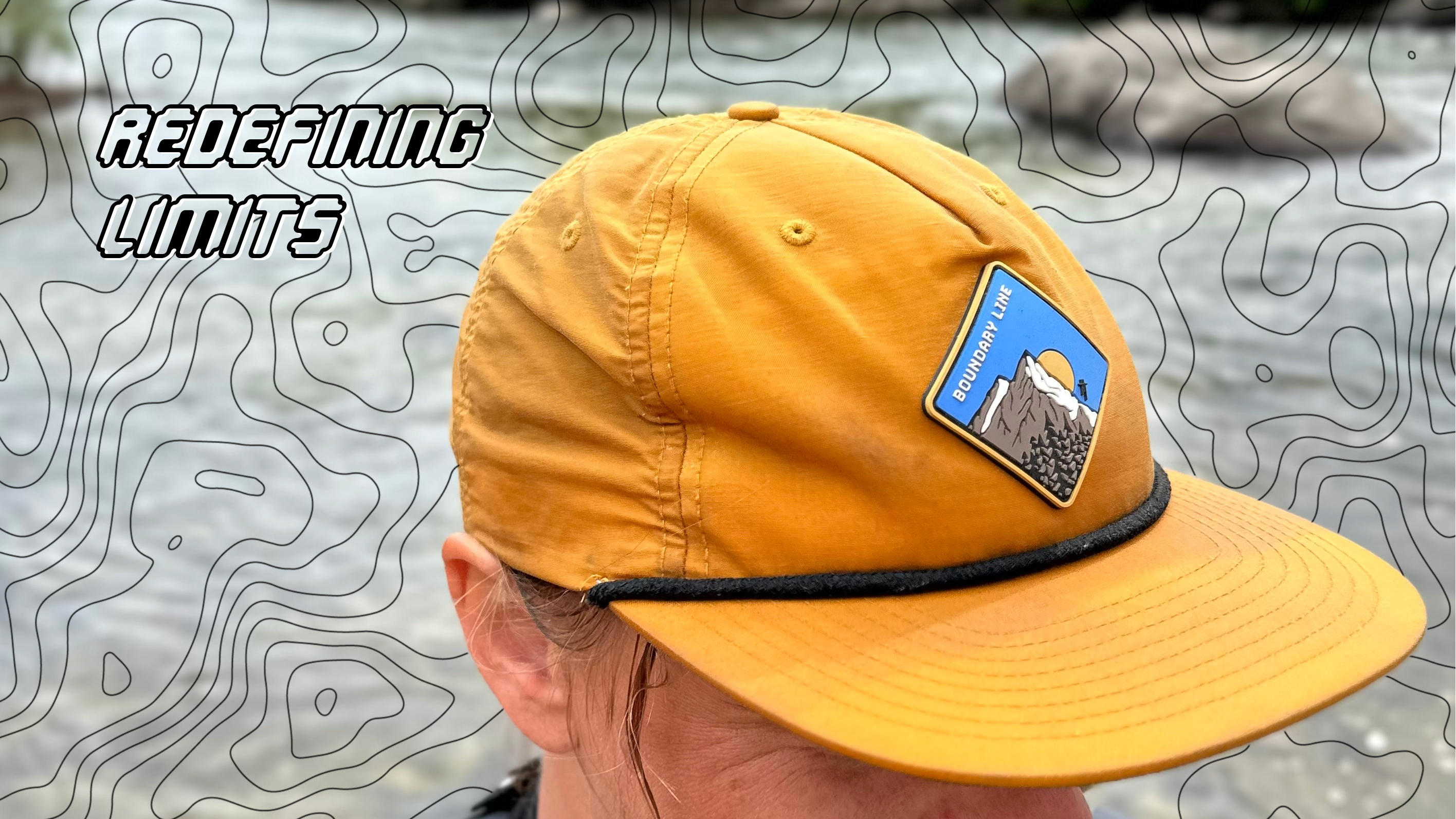 Person wearing a yellow cap with a patch, standing in front of a river with greenery.